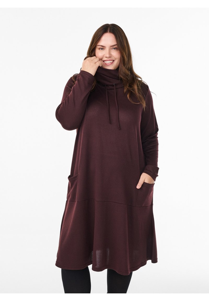 Donna che indossa un vestito bordeaux a maniche lunghe con collo a scialle e tasche, sorridente con una mano vicino al viso e l'altra nella tasca.