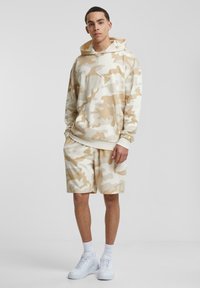 Beige kamuflasje hettegenser med frontlomme og matchende shorts. Har myk stoff med en avslappet passform. Kombinert med hvite joggesko.