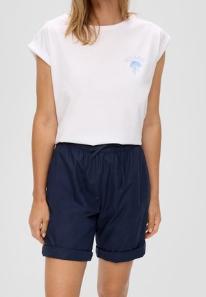 Kvinde iført en hvid kortærmet top med teksten "Voyage" og blåt blomsterdesign, kombineret med marineblå shorts med snøre og opsmøgede kanter.