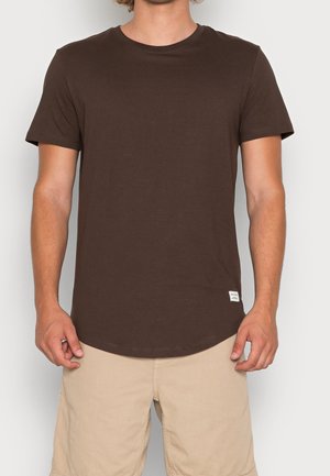 Homme portant un t-shirt uni marron foncé à manches courtes et un short beige clair, debout devant un fond clair uni.
