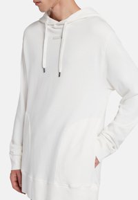 Felpa bianca con cappuccio realizzata in materiale morbido. Presenta una tasca frontale, cappuccio con lacci e una vestibilità leggermente oversize. Marchio visibile sul petto.