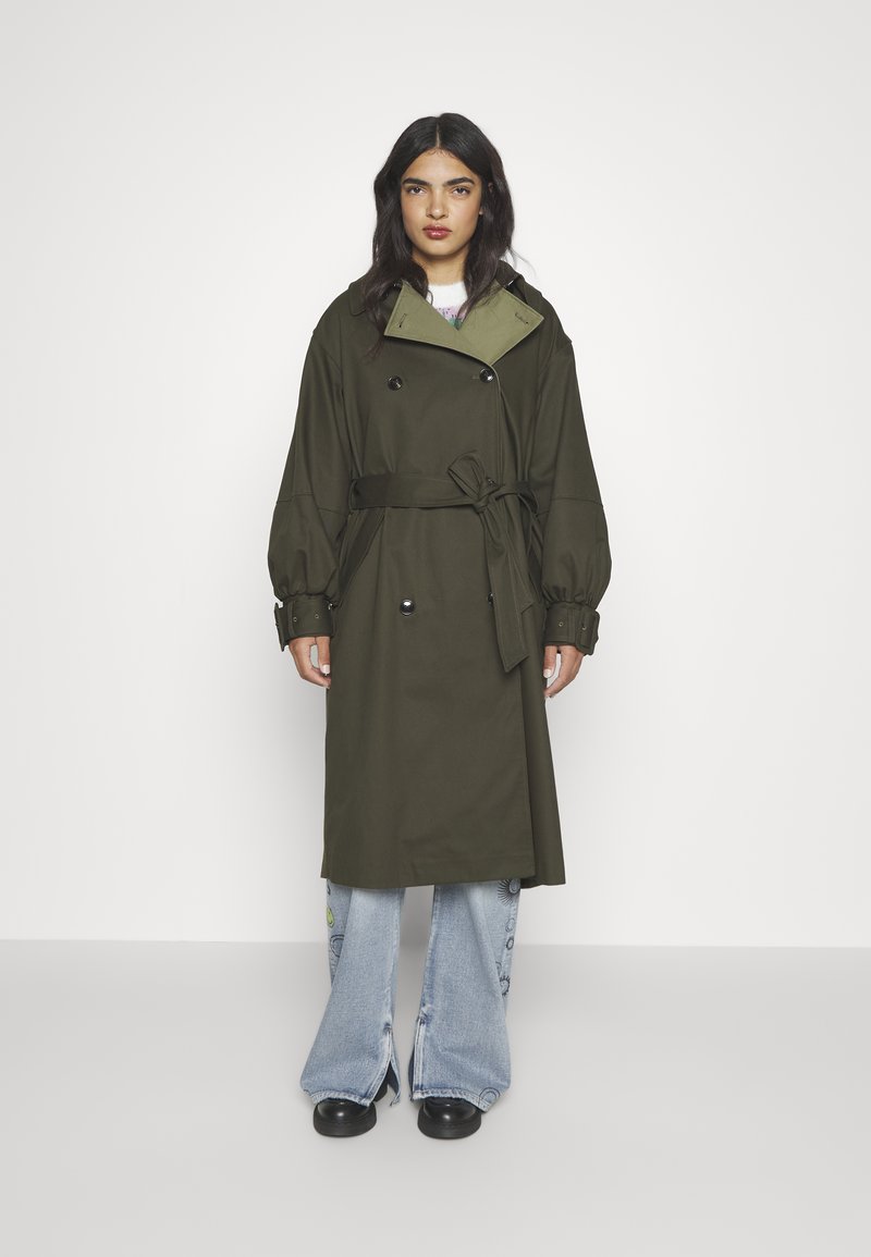 Olivgrüner Trenchcoat mit doppelter Knopfleiste, tailliertem Gürtel und übergroßen Ärmeln. Kombiniert mit hellblauen Schlagjeans und schwarzen Schuhen.