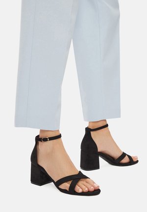 Pies con sandalias negras de ante, tacón bloque y tiras en el tobillo, combinadas con pantalones cortos de color claro sobre un fondo blanco.