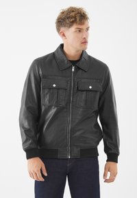Junger Mann in einer schwarzen Lederjacke mit zwei vorderen Klappen-Taschen und gerippten Bündchen, der vor einem weißen Hintergrund steht.