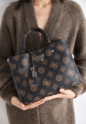 Person i brun sweater holder en sort Guess håndtaske med gyldne logo-mønstre og vedhæftet mærkat ved håndtagene tæt på brystet.