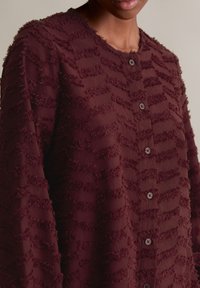 Bordowa bluzka z guzikami o teksturowanej powierzchni, z warstwowym, postrzępionym wzorem, okrągłym dekoltem oraz pięcioma guzikami pokrytymi tkaniną z przodu.