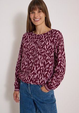 Femme souriante portant un chemisier bordeaux à manches longues avec un motif abstrait rose et un jean bleu, une main dans la poche, debout devant un fond uni.
