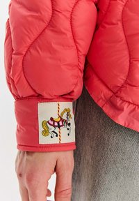 Rukáv červené prošívané bundy s nášivkou kolotočového koně, nošený přes světlé šedé džínové kalhoty na paži osoby.