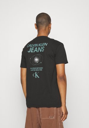 Mann trägt ein schwarzes Calvin Klein T-Shirt mit blauem, weltraumthemenbezogenem Motiv und Text auf der Rückseite, kombiniert mit brauner Hose.