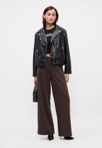 Μαύρο δερμάτινο μπουφάν μηχανής με ασημί αξεσουάρ, φορεμένο πάνω από ένα μαύρο crop top, συνδυασμένο με καφέ φαρδιά παντελόνια και μια μαύρη τσάντα με ακίδες.