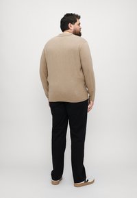 Beige dlouhý rukáv košile s límečkem, mírně texturovaná tkanina, v kombinaci s černými kalhotami a bílými teniskami. Pohled ze zádu.