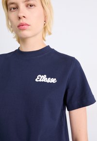 T-shirt en coton marine avec col rond, arborant un logo "Ellesse" blanc sur la poitrine gauche, des manches courtes et une texture lisse.