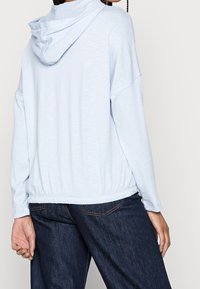 Ljusblå huvtröja i mjukt tyg, med avspänd passform, långa ärmar och elastisk nederkant. Designen inkluderar en framficka i känguru-stil.