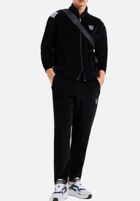 Tuta in velluto nero con giacca con zip, caratterizzata da strisce sulle spalle e un logo, abbinata a sneakers nelle tonalità di nero, grigio e blu.