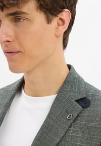 Grauer Blazer mit einem subtilen karierten Muster, ausgestattet mit einem schwarzen Akzent am Kragen und einem silbernen "D"-Logo. Getragen über einem weißen Hemd.