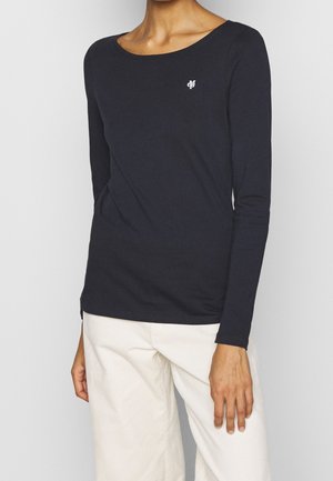 Persoon draagt een aansluitend zwart shirt met lange mouwen en een klein wit logo op de borst, gecombineerd met losse witte broek.