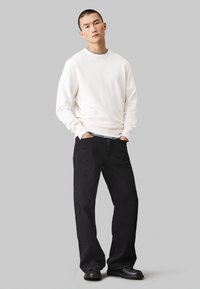 Felpa bianca con polsini a costine, abbinata a jeans neri a gamba larga. L'outfit è completato da stivali neri. Sfondo semplice.