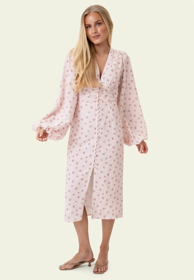 Femme blonde portant une longue robe fleurie rose avec des boutons sur le devant et des manches longues bouffantes, debout devant un fond neutre.