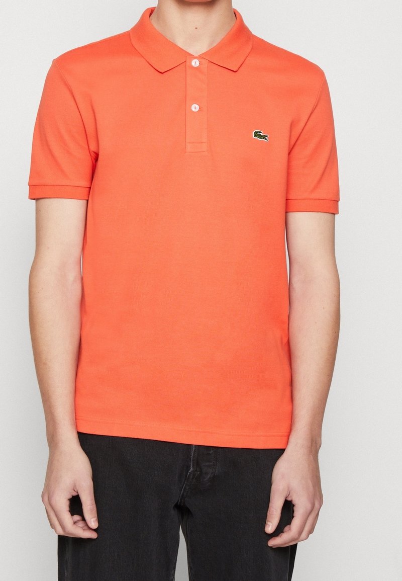 Homme portant un polo orange vif à manches courtes avec deux boutons blancs et un petit logo brodé sur la poitrine gauche, associé à un pantalon noir.