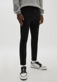 Personne portant un jean slim noir, un sweat gris, des chaussettes blanches et des baskets blanches avec des détails noirs, debout sur un fond clair.
