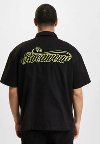 Fekete rövid ujjú póló, amelyen nagy sárga "Rocawear" logó és "est. '99" hímzés található a hátán; laza fazon, kényelmes illeszkedéssel.
