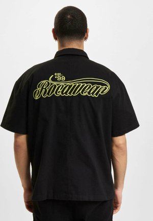 Fekete rövid ujjú póló, amelyen nagy sárga "Rocawear" logó és "est. '99" hímzés található a hátán; laza fazon, kényelmes illeszkedéssel.