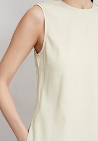 Ärmelloses beiges Kleid mit glatter Textur, minimalen Nähten, einem geschwungenen Ausschnitt und seitlichen Taschen, die in das Design integriert sind.