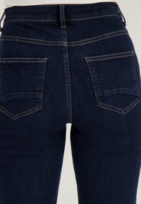 Donkerblauwe spijkerbroek van achteren gezien, met twee achterzakken met zichtbare oranje stiksels.