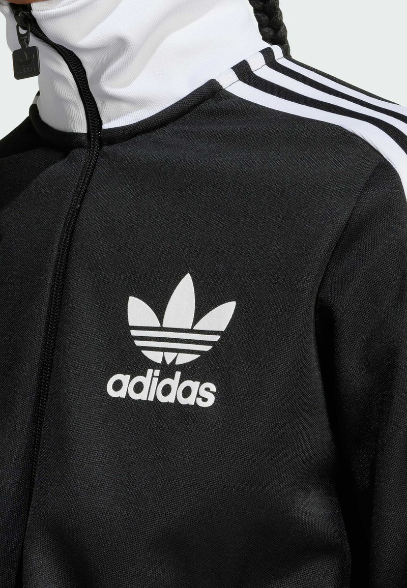 Chaqueta de pista negra con un cuello alto, cremallera blanca y tres rayas blancas en los hombros. Presenta un gran logo blanco de Adidas en el pecho.