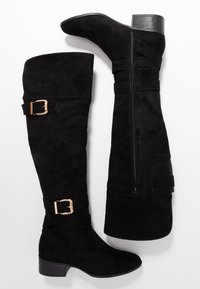 Botas negras de ante por encima de la rodilla, con cremallera lateral, correas anchas con hebillas en tono dorado y tacón ancho.