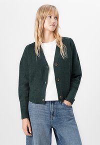 Kvinne med langt blondt hår ikledd en mørk grønn knappet strikkegenser, hvit bluse med volanger i kragen, og blå jeans, som ser til siden.