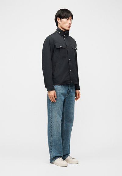 Νεαρός άνδρας που στέκεται πλαγιαστά φορώντας μαύρο σακάκι με κουμπιά, ανοιχτόχρωμα τζιν και λευκά αθλητικά παπούτσια σε απλό φωτεινό φόντο.