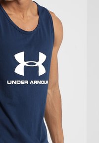 Laivastonsininen, hihaton tankkitoppi, valmistettu puuvillasta, jossa suuri valkoinen Under Armour -logo edessä. Sileä pinta, perusmuotoilu.