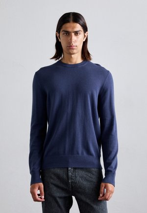 Jeune homme avec des cheveux bruns mi-longs portant un pull bleu marine à manches longues et un pantalon sombre, se tenant contre un fond gris uni.