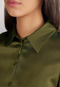 Blusa di seta verde scuro con colletto a punta, dotata di una chiusura frontale a bottoni e una superficie liscia e lucida.