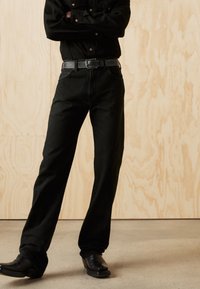 Zwarte denim broek met een rechte snit, gecombineerd met een zwarte riem met een zilveren gesp, gedragen met zwarte schoenen tegen een lichte houten achtergrond.