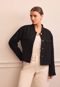 Giacca nera texturizzata con collo rotondo, tasche frontali e chiusura con bottoni, abbinata a pantaloni a vita alta di colore chiaro.