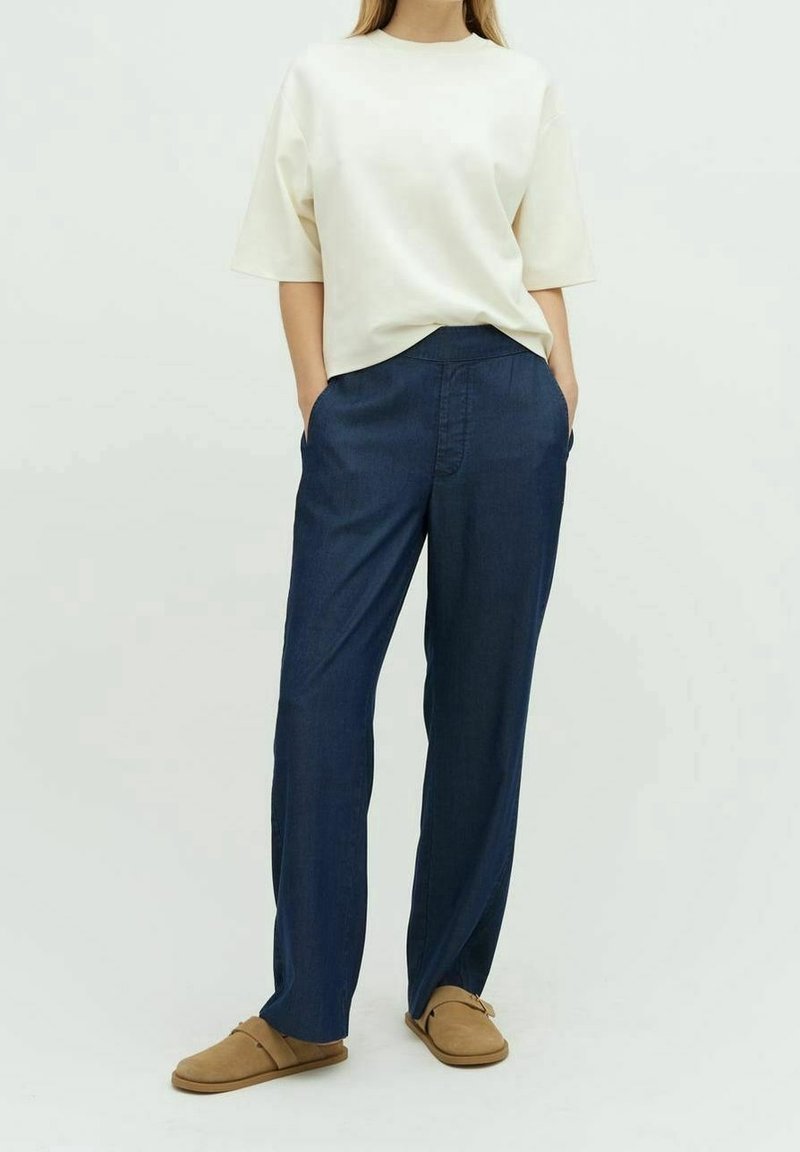 Femme portant un haut blanc ample à manches courtes, un pantalon large bleu foncé, et des chaussures plates marron clair, debout les mains dans les poches sur un fond uni.