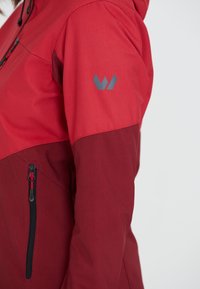 Röd och vinröd jacka med en dragkedjeficka, justerbar huva och en grå logotyp på axeln, tillverkad av ett slätt, tekniskt tyg.