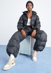 Černá péřová bunda s kapucí, v kombinaci s volnými černými péřovými kalhotami. Bílý crop top a béžové sportovní boty s černými detaily.
