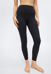 Γυναίκα που φοράει μαύρο κολάν ψηλόμεσο, όρθια και ξυπόλητη μπροστά σε απλό ανοιχτό φόντο, με τα χέρια χαλαρά δίπλα στο σώμα.