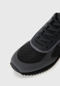 Black athletic shoe with mesh and synthetic materials; features a textured surface, flat laces, and a rubber outsole with a tread pattern.