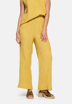 Mujer con pantalones anchos texturizados color mostaza y top sin mangas a juego, con sandalias de tacón verde oliva.