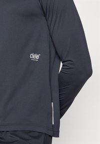 Camisola atlética de mangas longas em cinza escuro, feita de um tecido leve com detalhe de logotipo. Apresenta uma textura suave e acabamentos refletivos.