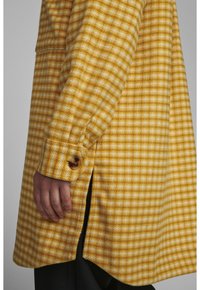 Žlutý a bílý kostkovaný kabát s texturovanou látkou, knoflíkovým manžetou a bočními rozparky pro zvýšenou pohyblivost.