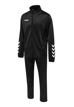 Mann, der die Manschette einer schwarzen Sportjacke mit weißem Pfeildesign sowie passenden schwarzen Sporthosen und Schuhen vor grauem Hintergrund anpasst.