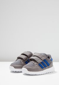 Harmaat urheilukengät sinisin raidoin, joissa on tarrakiinnitys ja teksturoitu kangaspäällinen. Valkoiset kumipohjat tarjoavat pidon.