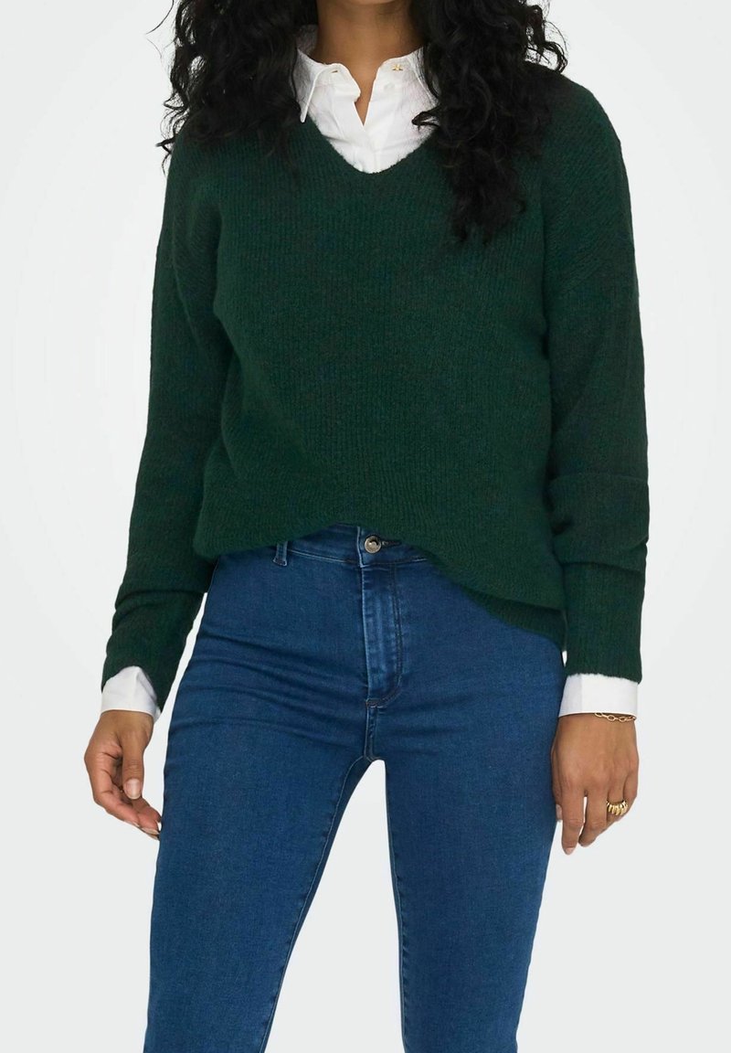Femme portant un pull vert foncé à col en V sur une chemise blanche, associé à un jean bleu taille haute, debout devant un fond uni.