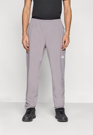 Homme portant un pantalon The North Face gris clair avec des chaussures noires et un t-shirt noir, debout sur un fond blanc uni.