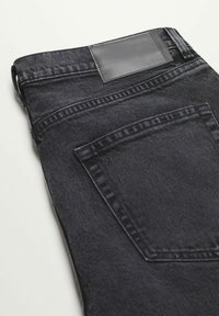 Jeans de mezclilla negra con un parche de cuero en la cintura, que cuentan con un único bolsillo trasero y detalles de costura visibles en una tela texturizada.